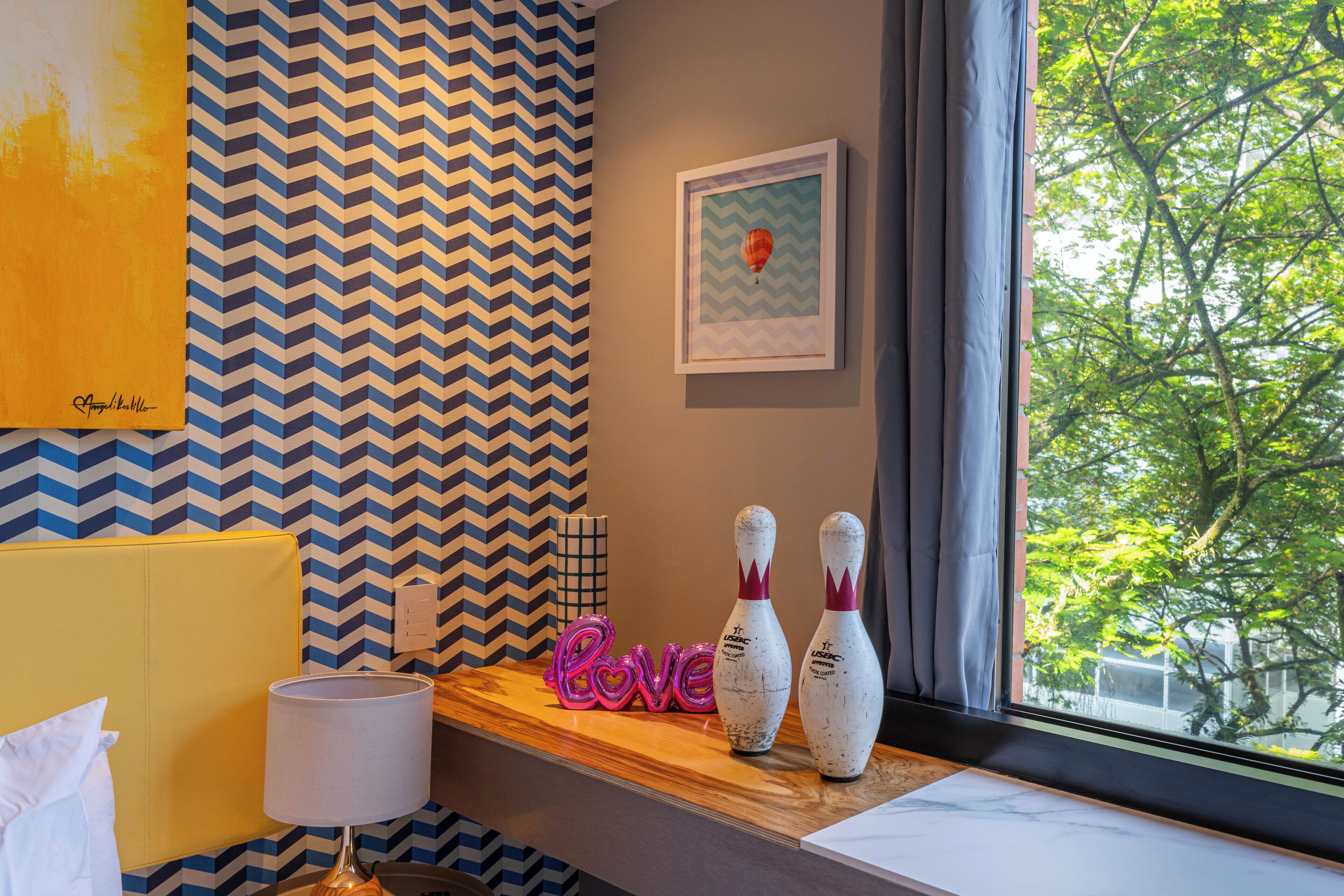 A hotel room with a chevron-patterned wall, a window with a view of trees, a wooden shelf with bowling pins and a 'Love' sign, and a lamp on a bedside table.