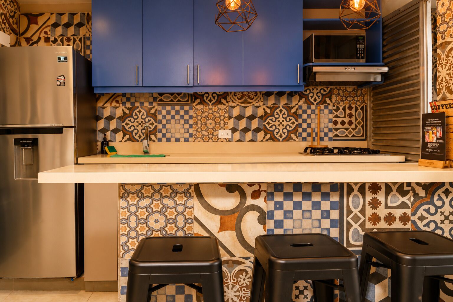 A kitchen with blue cabinets, a stainless steel refrigerator, a microwave, and a gas stove. There are two black stools and a green cutting board on the counter.