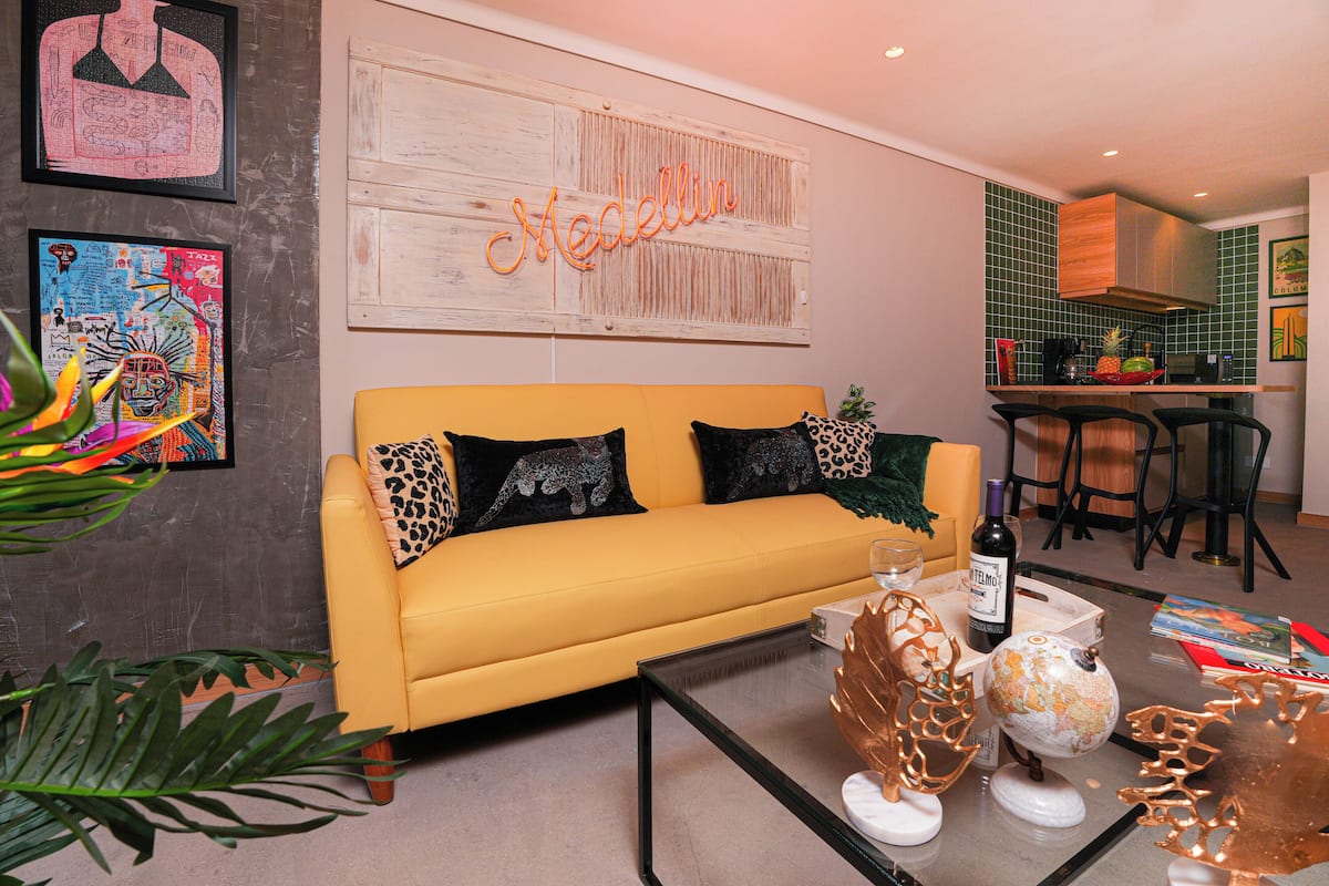 A living room with a yellow sofa, a glass coffee table, and a kitchen area with a bar.
