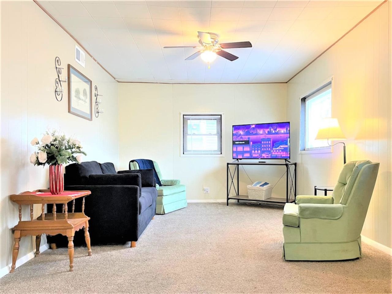 A living room with a ceiling fan, a television, a green armchair, a sofa, and a small table with a vase of flowers.
