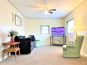 Un salon avec un ventilateur de plafond, un téléviseur, un fauteuil vert, un canapé et une petite table sur laquelle est posé un vase de fleurs.