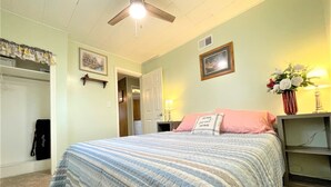 Un dormitorio con cama, ventilador de techo, mesita de noche, lámpara y estante en la pared.