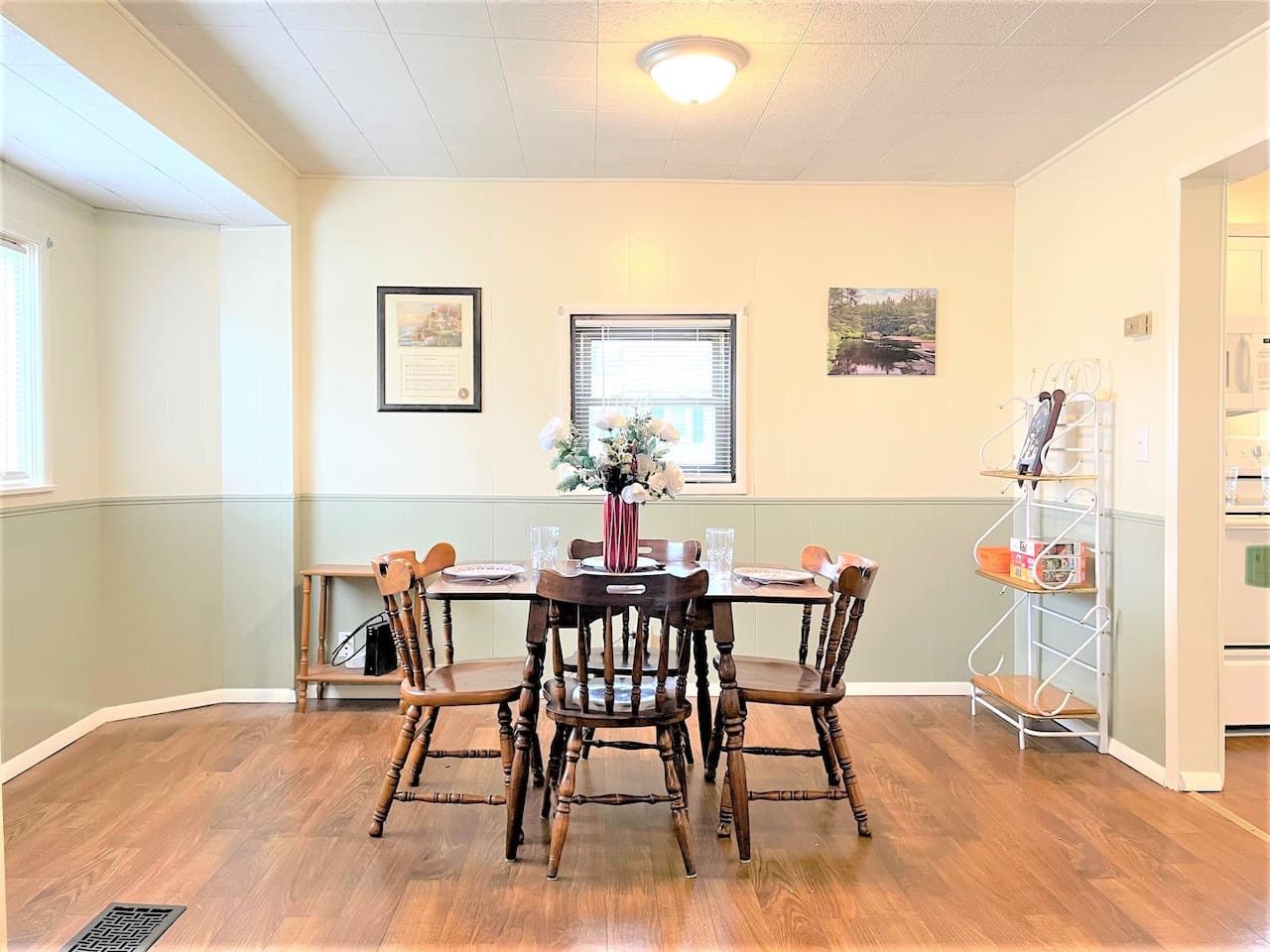 A dining area with a wooden table and chairs, a vase with flowers, framed pictures on the wall, and a window with blinds.