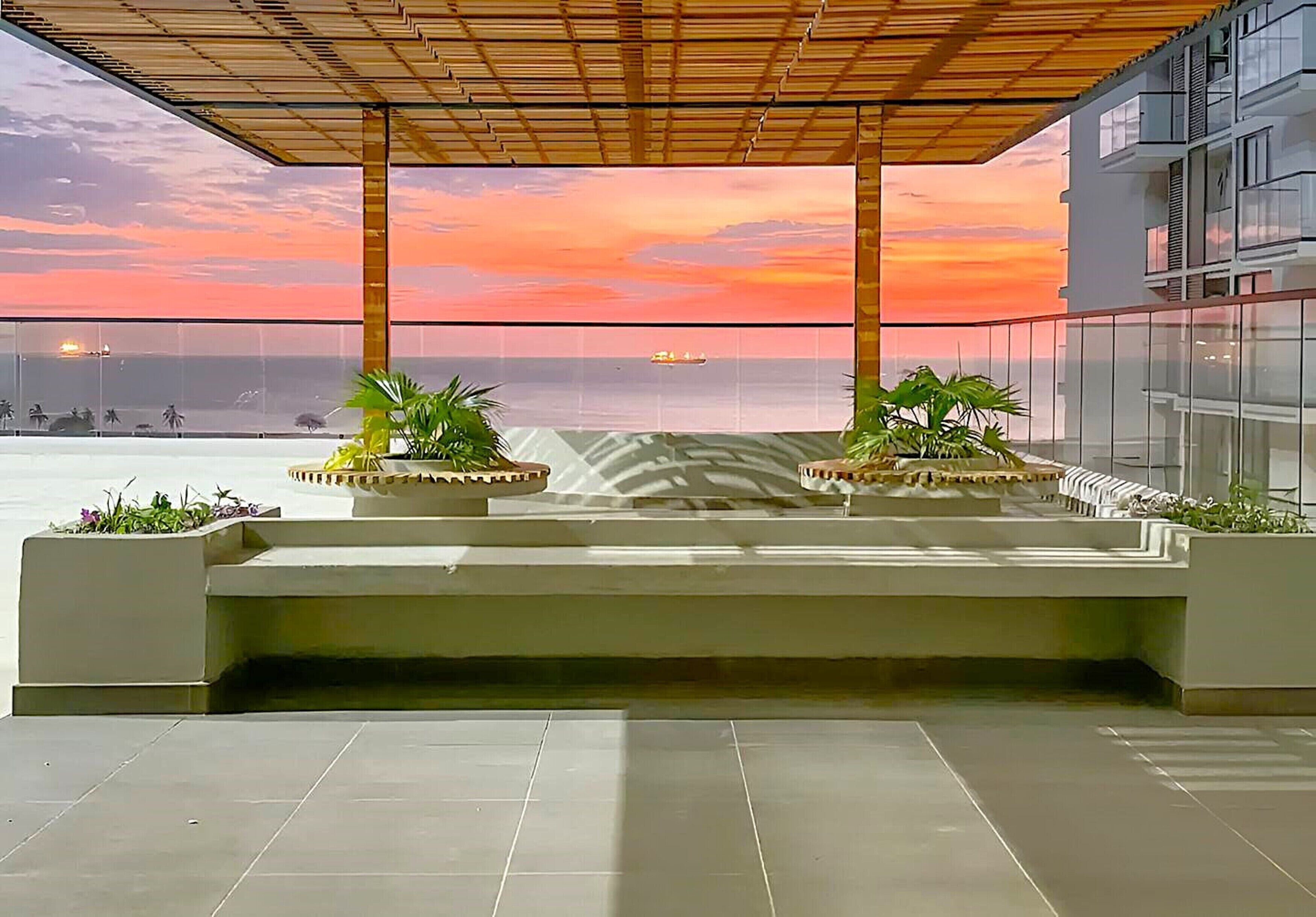 A rooftop terrace with a view of the sunset over the ocean, featuring two planters with plants and a modern architectural design.