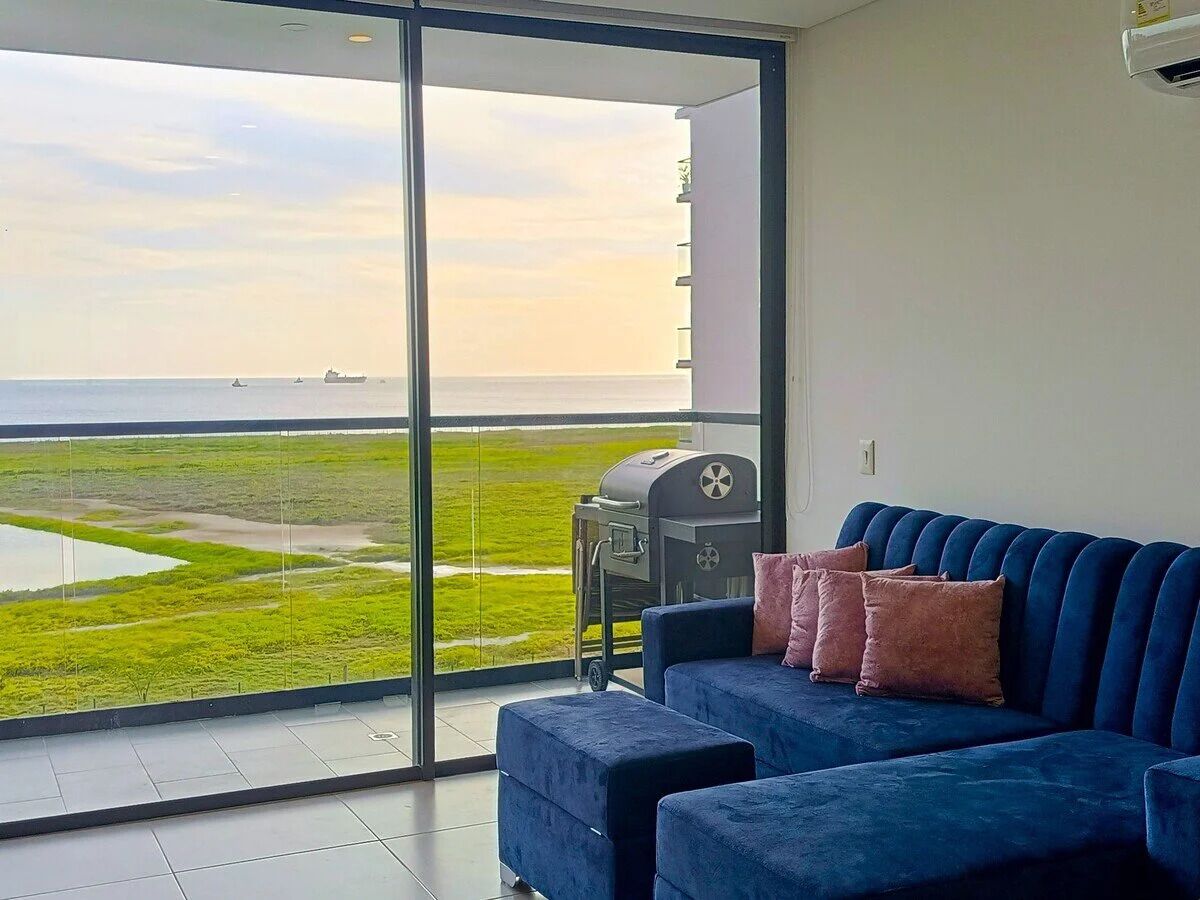 A balcony with a view of a green landscape and the sea, a blue sofa with cushions, and a barbecue grill.