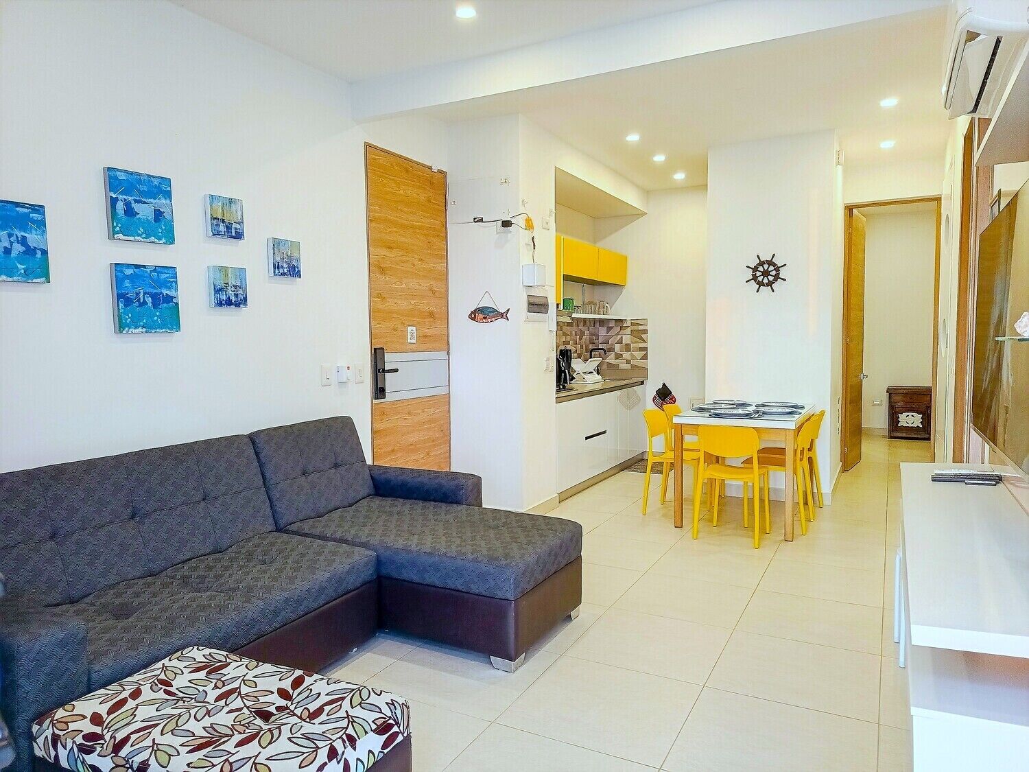 A modern living room with a grey sectional sofa, a small patterned ottoman, and a dining area with yellow chairs and a white table.