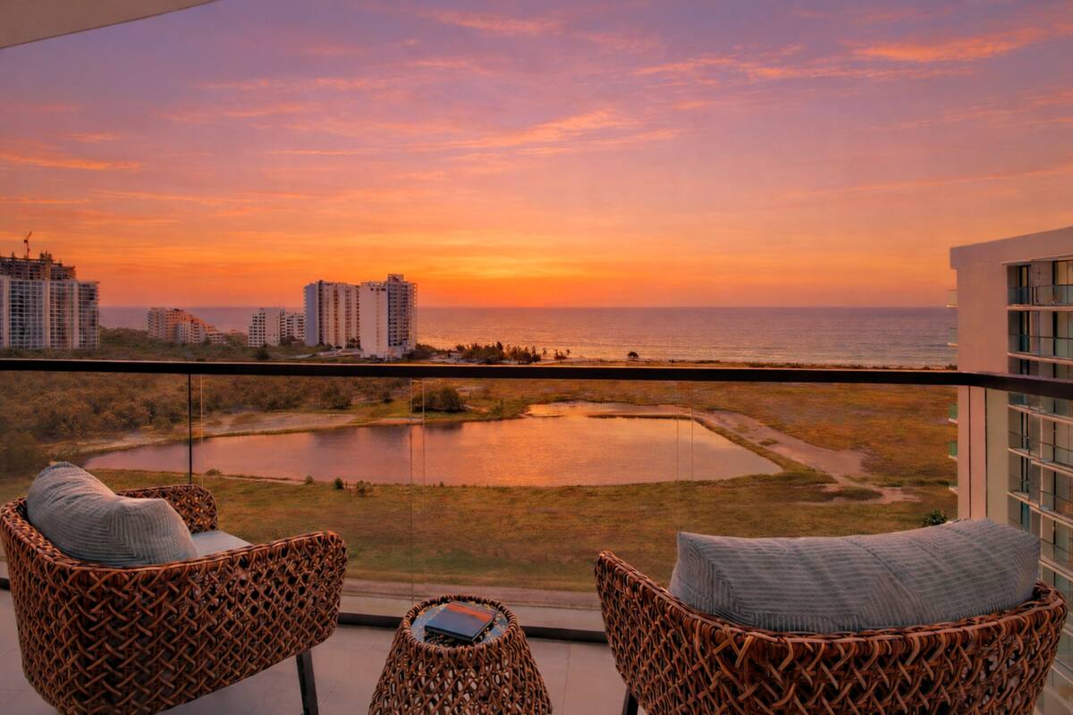 A balcony with wicker chairs and a view of a sunset over a body of water and buildings.