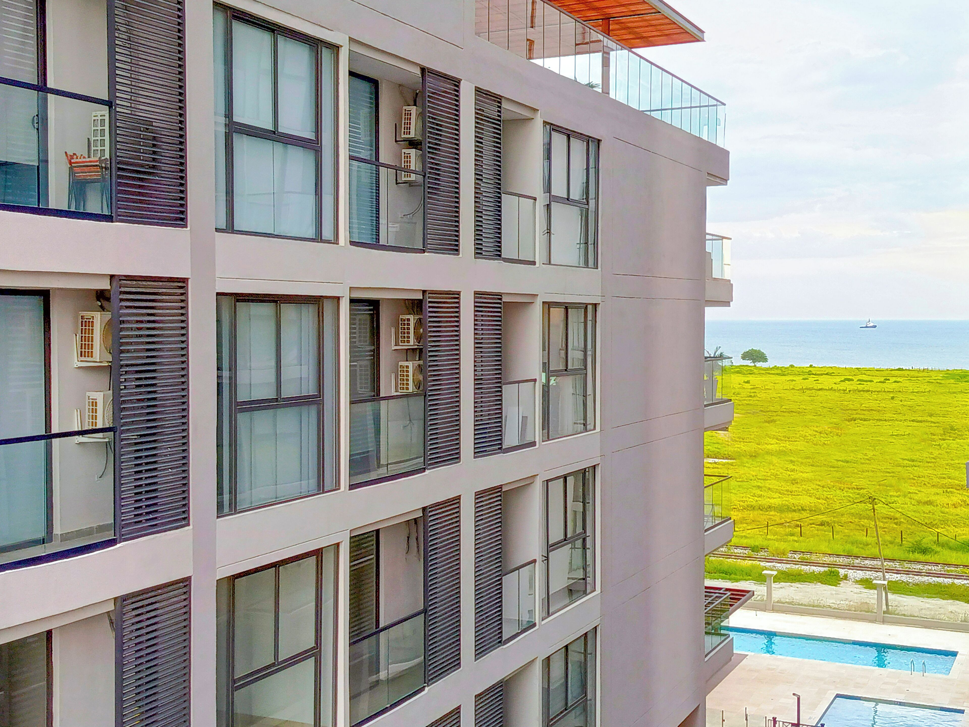 Un edificio de varios pisos con ventanas amplias y unidades de aire acondicionado. Se observa un campo verde y una masa de agua al fondo.