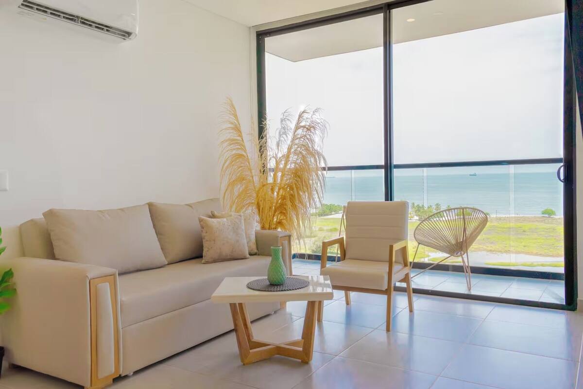 A modern living room with a sofa, a small table, and a chair. There is a large window with a view of the sea.