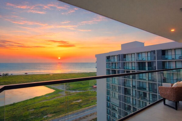 A balcony with a view of the sunset over the ocean, a modern building with large windows, and a chair.