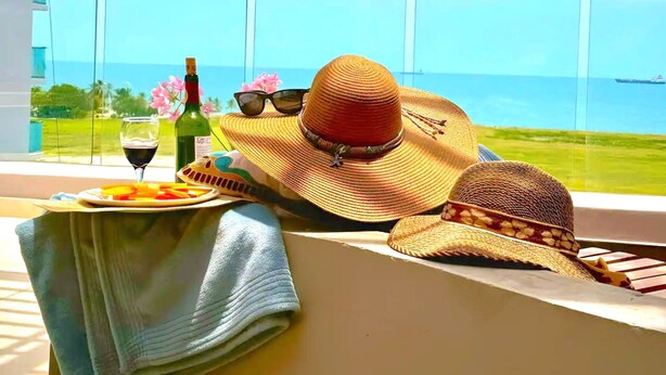 Een balkon met uitzicht op de oceaan, een fles wijn, een bord fruit en een strohoed.