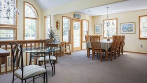 A dining area with a glass table, wooden chairs, and a chandelier. There are large windows with curtains, a door, and framed pictures on the walls.