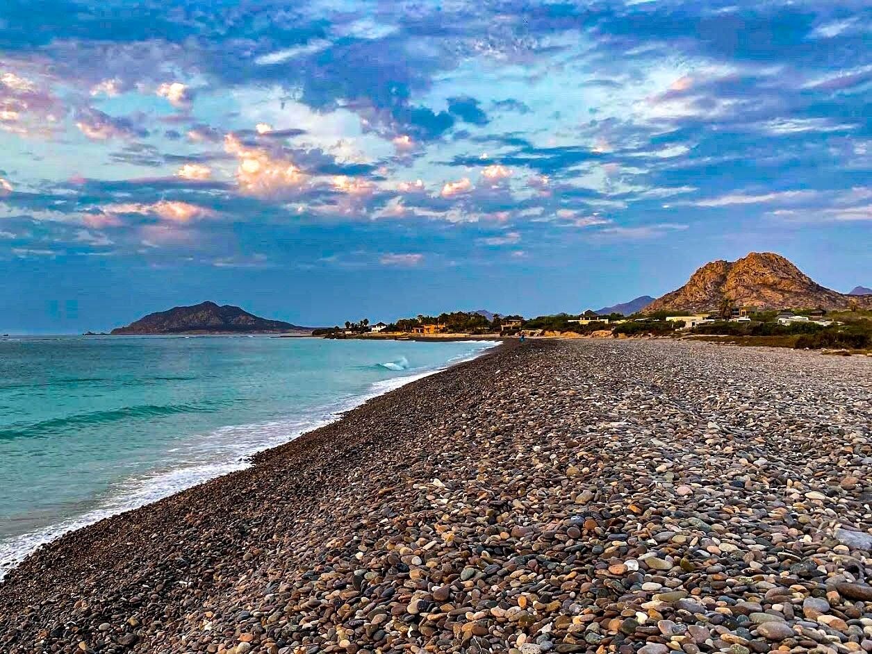 Ein Kiesstrand mit einem Berg im Hintergrund und blauem Himmel.