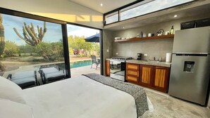 Ein modernes Schlafzimmer mit einem großen Bett, einer Küchenzeile mit Kühlschrank und Blick auf Pool und Garten.