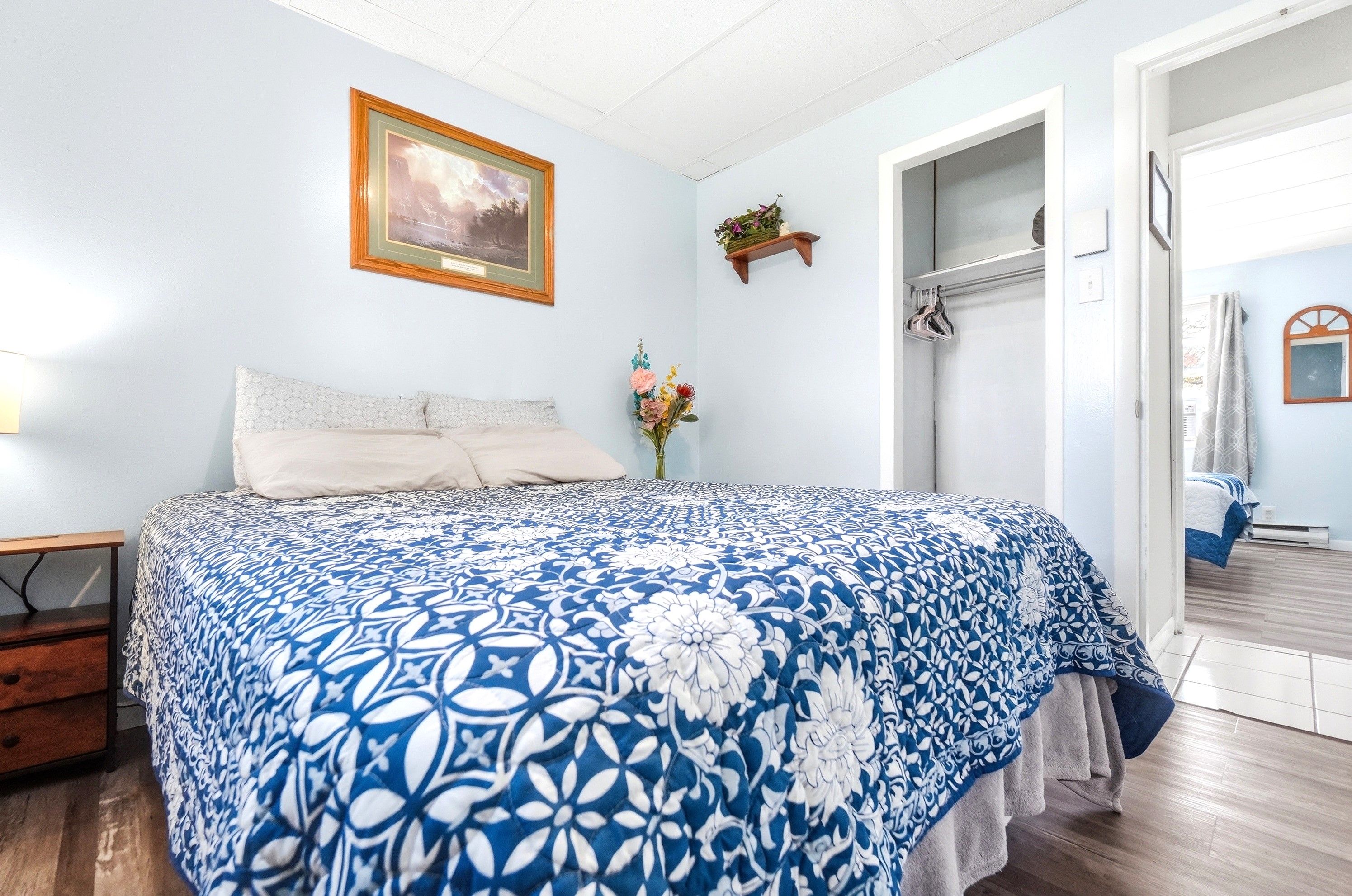 A bedroom with a bed, a nightstand, a wall-mounted shelf with flowers, and a door leading to another room.
