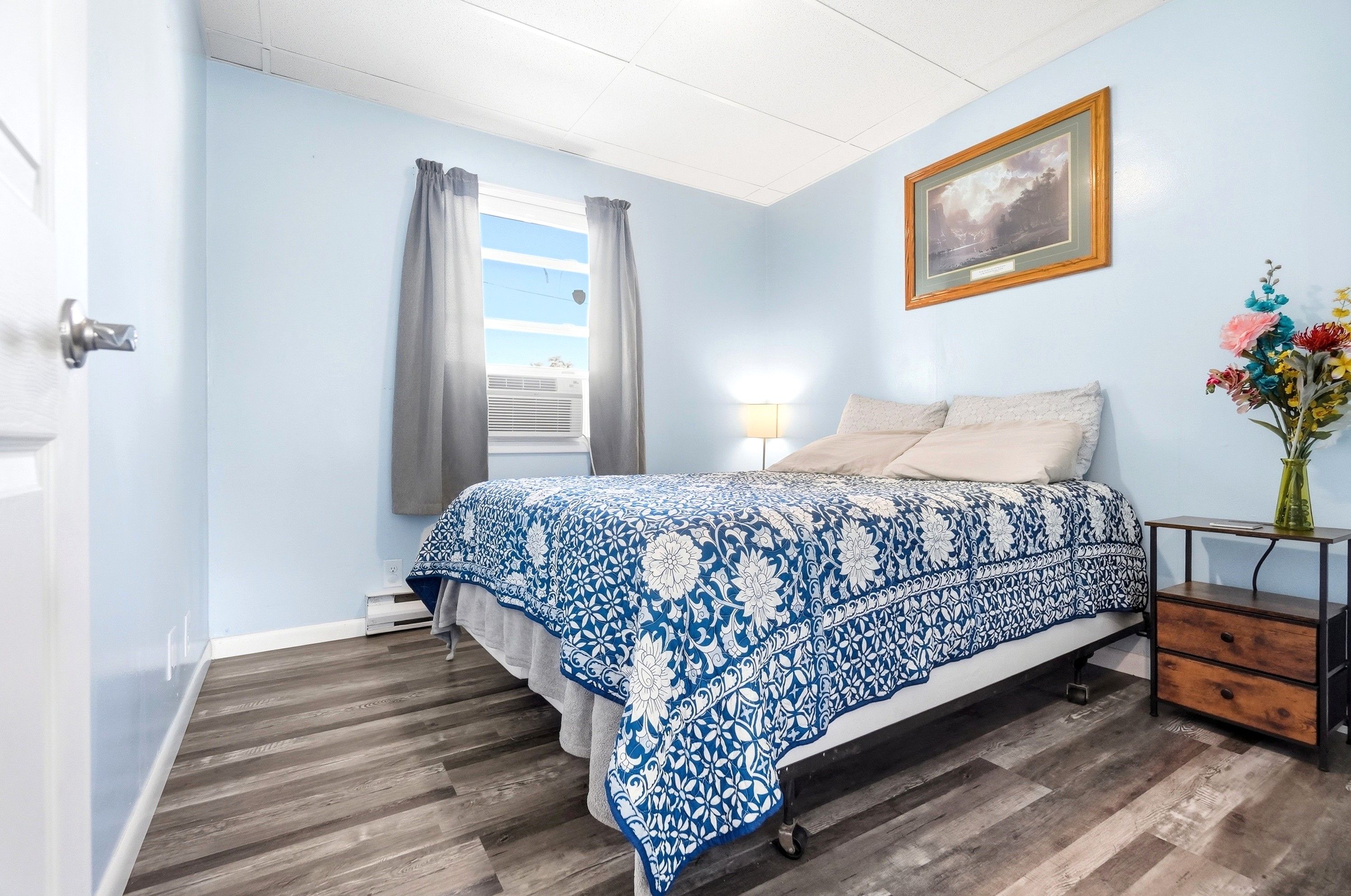 Une chambre à coucher avec un lit, une table de chevet, une lampe, une fenêtre avec des rideaux, un tableau au mur et un vase avec des fleurs.
