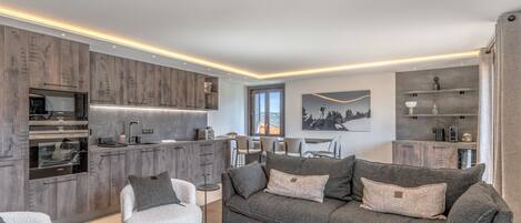 Un salon moderne avec un canapé gris, un fauteuil blanc et une table basse. On aperçoit, au fond, une cuisine avec des meubles en bois et un four encastré.