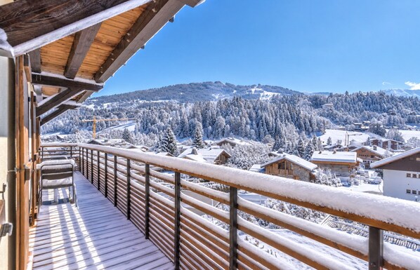 A wooden balcony with a metal railing overlooking a snowy landscape with houses and trees.