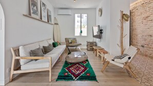 A modern living room with a white sofa, a coffee table, and a colorful rug. There is a surfboard on the wall, a TV, and a brick wall. - Maison Roccia - Welkeys (Anglet)