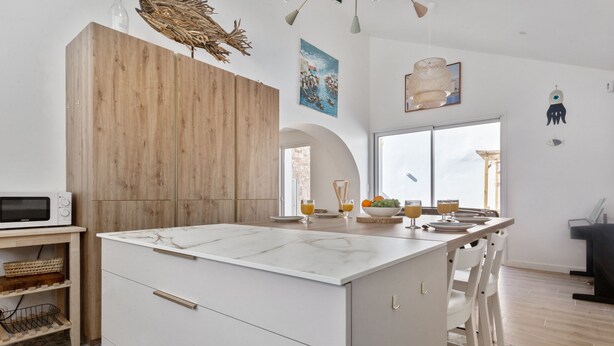 A modern kitchen with a central island, wooden cabinets, a microwave, and a dining area with a view. - Maison Roccia - Welkeys (Anglet)
