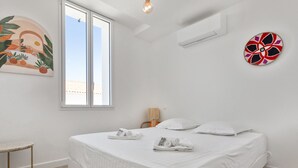 A bedroom with a large bed, a small round table, a wall-mounted air conditioner, and a window with a view of buildings. - Maison Roccia - Welkeys (Anglet)