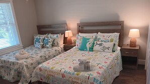 Ein Schlafzimmer mit zwei Betten, jeweils mit einem Holzkopfstück, einer Nachttischlampe und einer Tagesdecke im Strandstil.