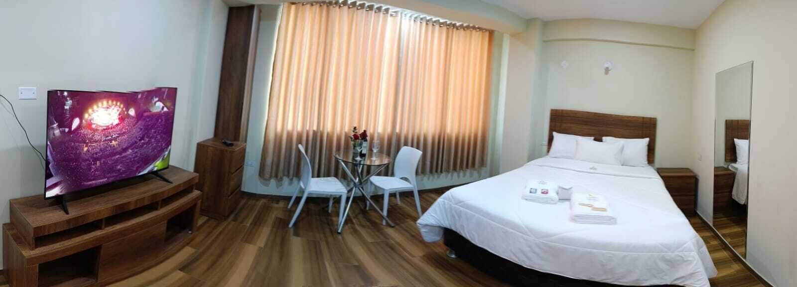 A hotel room with a bed, a TV, a small table with chairs, and a window with curtains.