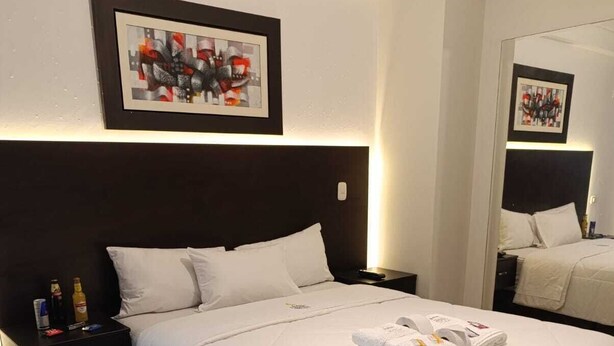 A neatly made bed with white bedding and a folded towel on top. There is a nightstand with a bottle and a remote control beside the bed. A framed picture hangs above the bed. - Hotel Arabian (Lima)