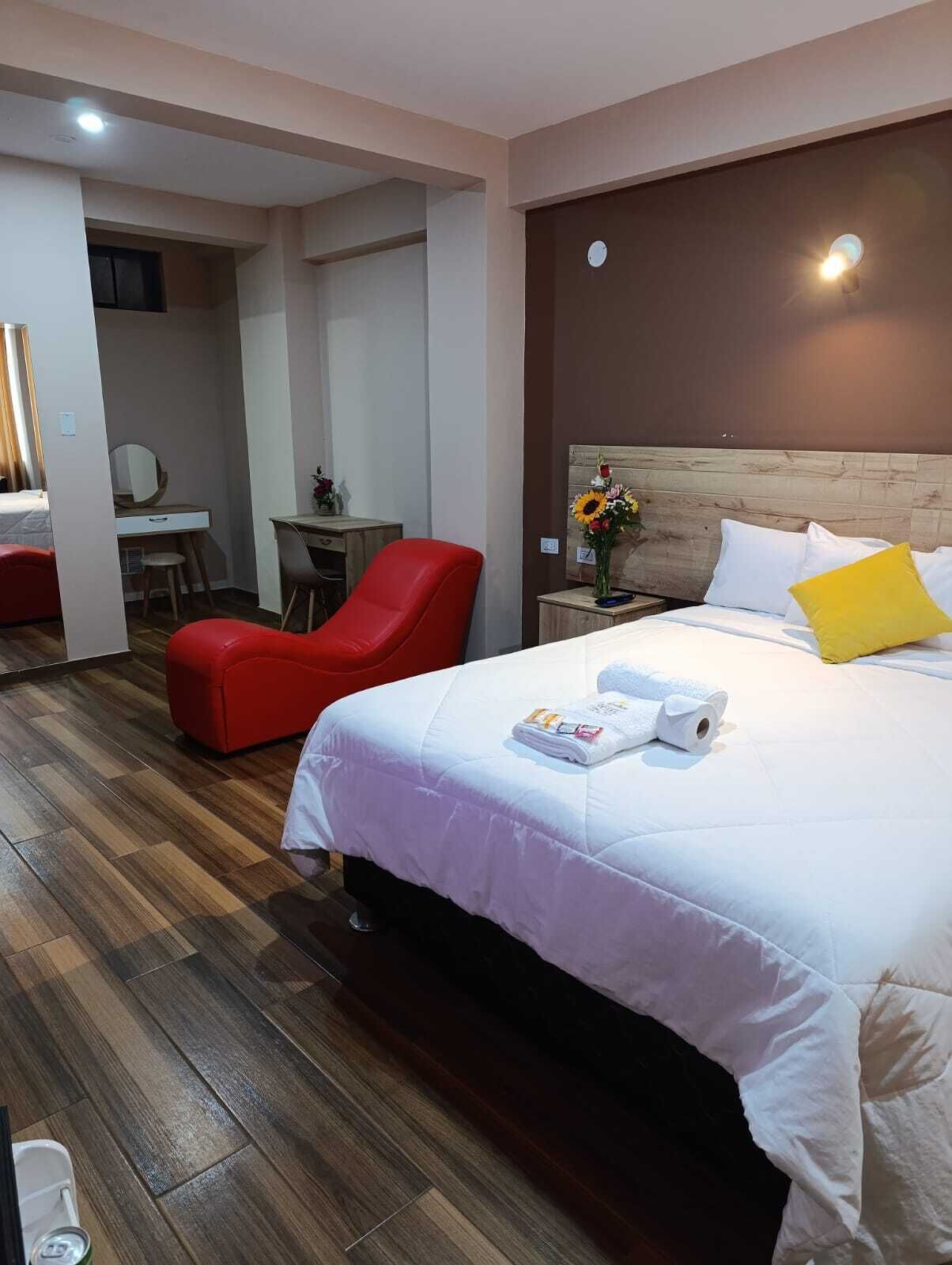 A hotel room with a bed, a red chair, a desk, and a small table with flowers.