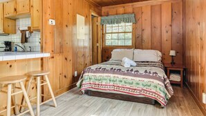 Interior de una cabaña de madera con una cama, una pequeña kitchenette y un ventanal.