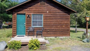 Una cabaña de madera pequeña con una puerta verde, una ventana y dos sillas afuera.
