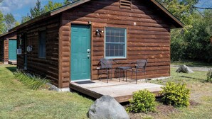 Una cabaña de troncos con puerta verde, ventana y un patio con sillas.