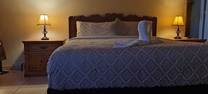 Una cama bien hecha con una colcha estampada, dos lámparas de noche y una mesita de noche de madera con un cajón.