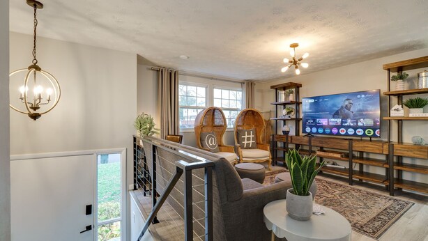 A modern living room with a large flat-screen TV, wicker chairs, a chandelier, and a staircase leading to an outdoor area. - Game Room-Deck-Coffee Bar (Englewood)