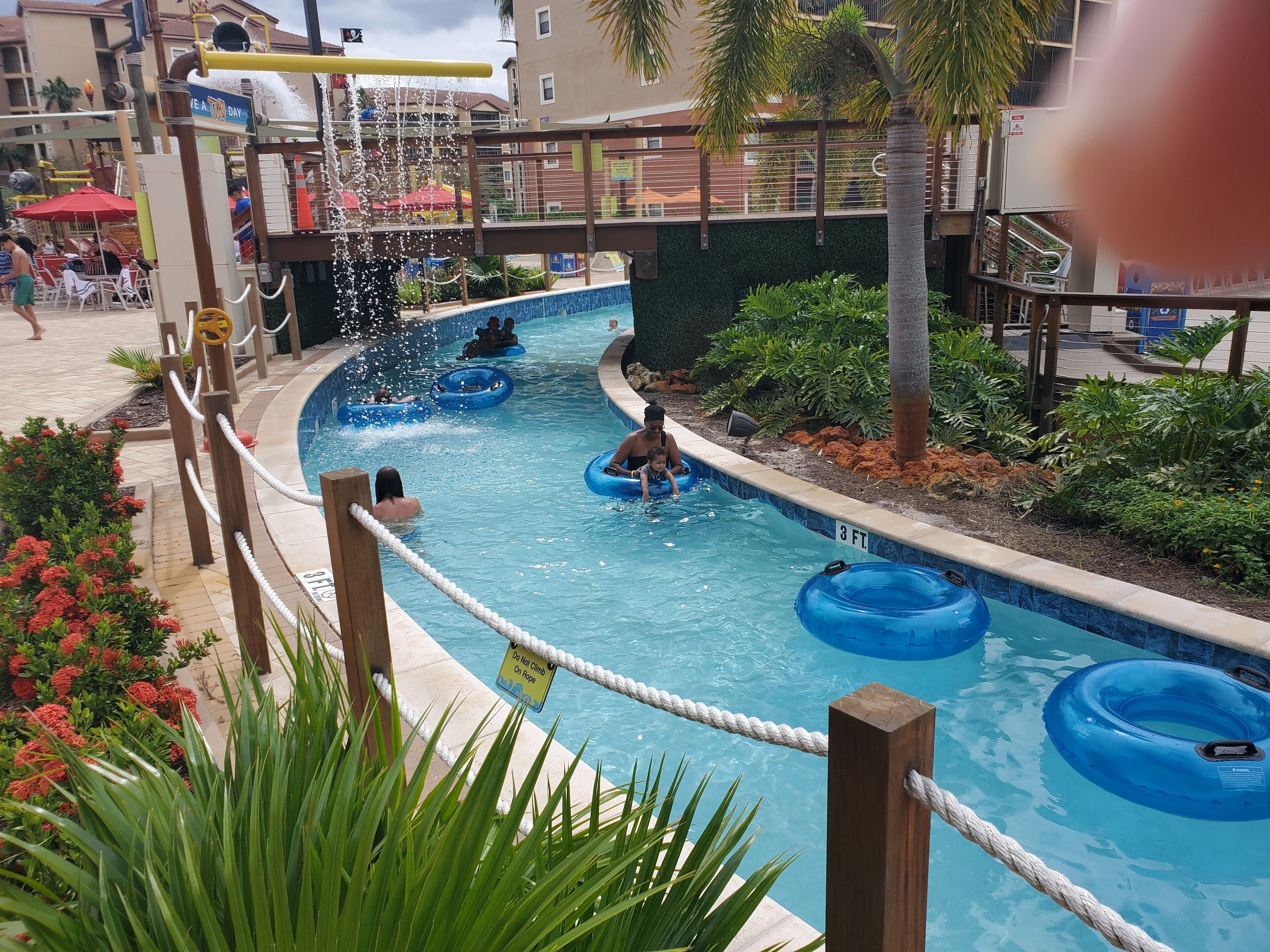A pool area with water slides and inflatable rings, surrounded by palm trees and a poolside area with chairs and umbrellas.