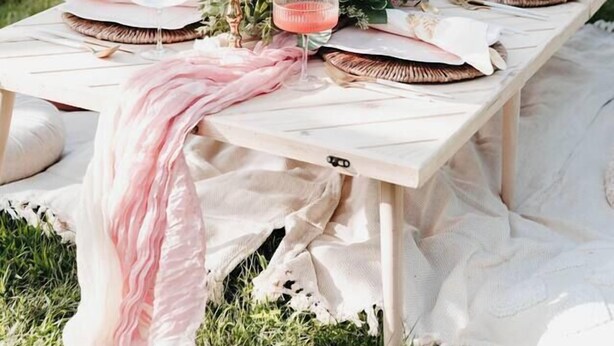 Una mesa de picnic dispuesta sobre césped, con un centro de mesa floral, servilletas blancas y detalles en rosa.