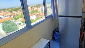 A room with a large window, a bench, a stainless steel refrigerator, and a view of a residential area. - Appartement T2 vue Mer, Moderne, Clim 3 Etoiles Parking 4plmer53 (CANET PLAGE)
