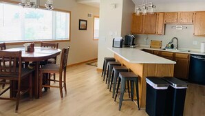 A modern kitchen with wooden cabinets, a central island with stools, a dining table with chairs, and a large window providing natural light.