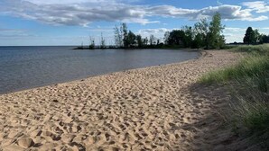 A sandy beach with a calm lake, surrounded by trees and a partly cloudy sky.