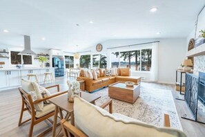 Amplia sala de estar con un sofá seccional grande, sillas de madera y una mesa de centro. Se observa una cocina al fondo con armarios blancos y un ventanal.
