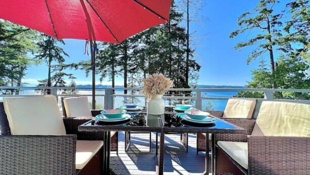 Un área de comedor al aire libre con mobiliario de mimbre, una mesa dispuesta con platos y un florero, y una sombrilla roja que proporciona sombra.