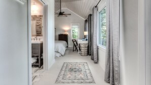 A narrow hallway leading to a bedroom with a bed, a desk, and a chair. There is a window with curtains, a ceiling fan, and a rug on the floor.