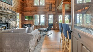 A cozy log cabin interior with a stone fireplace, comfortable seating, and large windows offering views of the outdoors.
