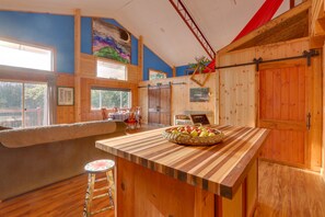 A kitchen with wooden cabinets, a large island with a basket of fruit, and a view of the outdoors through large windows. - Peaceful Berkeley Springs Home: Escape to Nature! (Berkeley Springs)