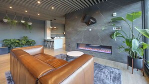 A modern lounge area with a leather sofa, hanging planters, a fireplace, and a kitchen in the background.