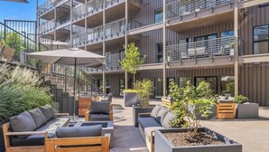 A modern outdoor seating area with wooden furniture and planters, a staircase leading to an upper floor, and a multi-story building with balconies.