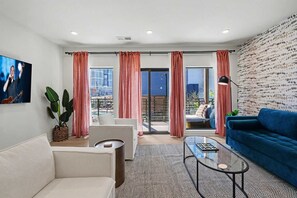 A modern living room with a sofa, a coffee table, and a blue couch. There is a large window with pink curtains, a plant, and a city view.