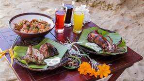 Une table dressée avec des viandes grillées, un saladier de crevettes et des boissons, le tout dans un cadre de plage.