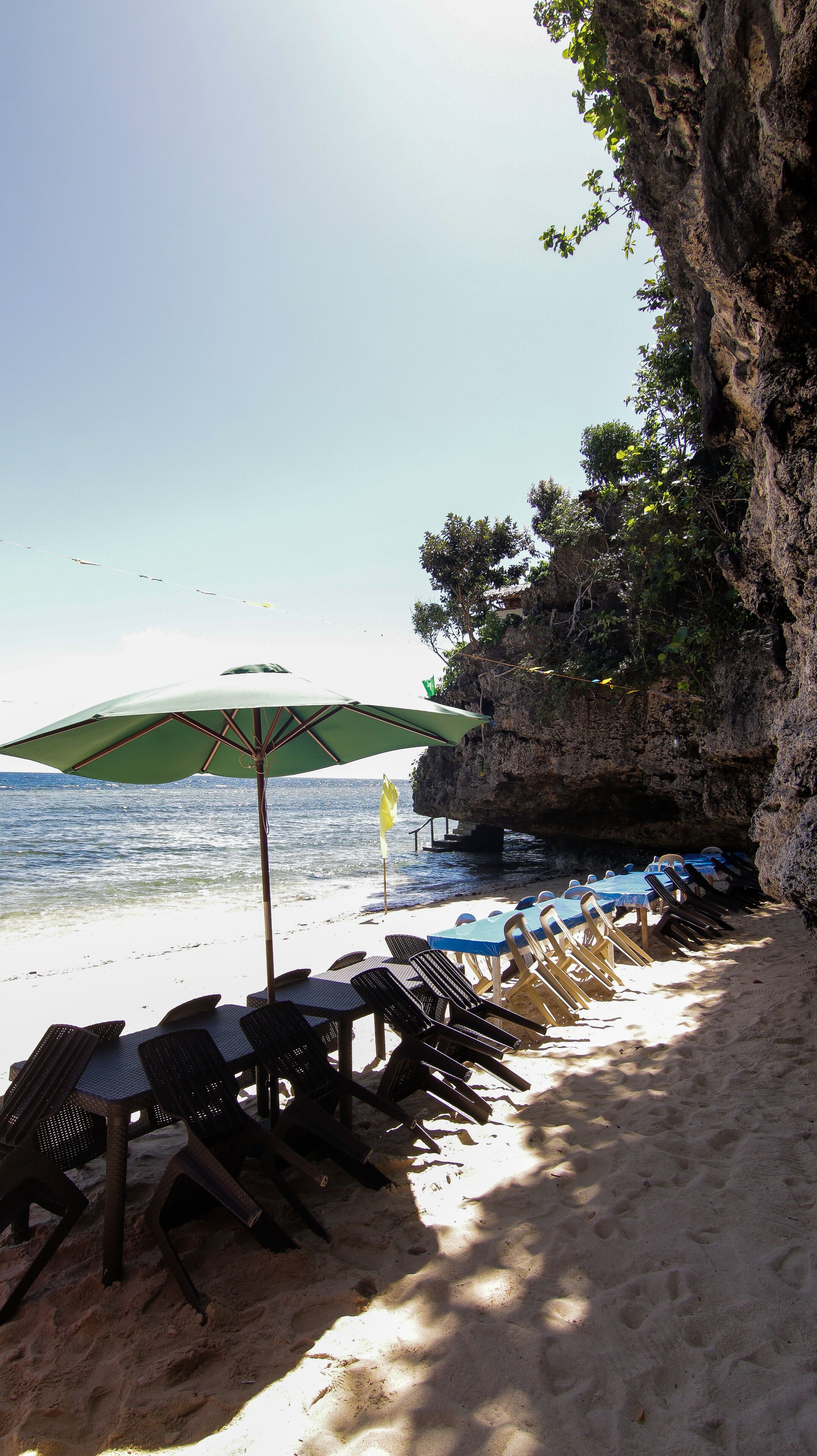 A beach with rows of sun loungers and a large umbrella, a rocky cliff, and the ocean in the background.