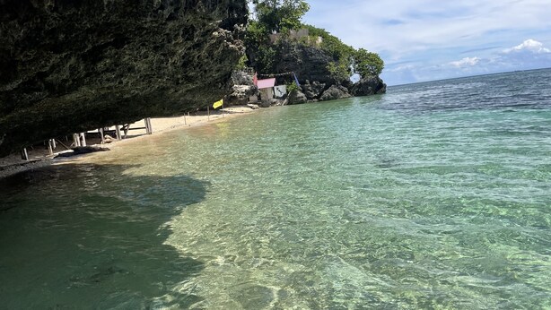 Una playa con agua cristalina, una pequeña cabaña y un acantilado rocoso.